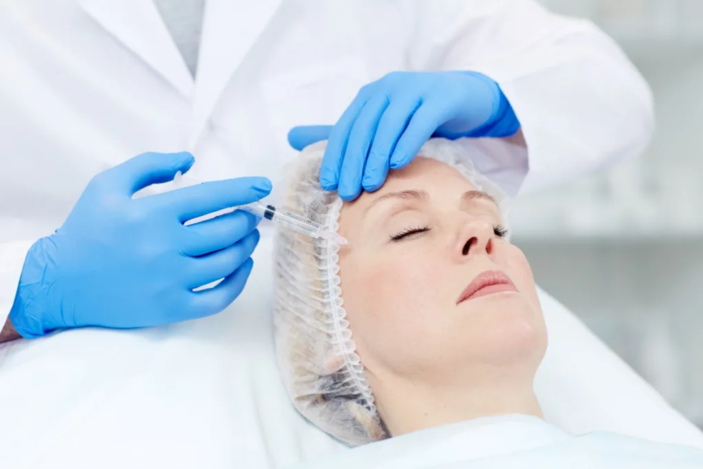 A serene woman with her eyes closed, wearing a hair net, receives an injection in her forehead from a medical professional wearing blue gloves and a white lab coat. This treatment is often considered the best solution for wrinkles by many.