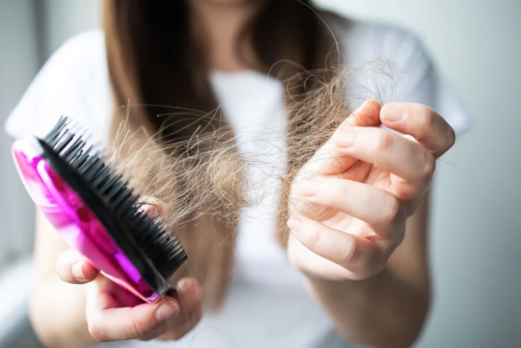 A close-up of a person with long brown hair, holding a pink brush in one hand and a clump of hair they have brushed out in the other, suggesting hair loss, as they look into the subject of hair loss treatment in Novi.