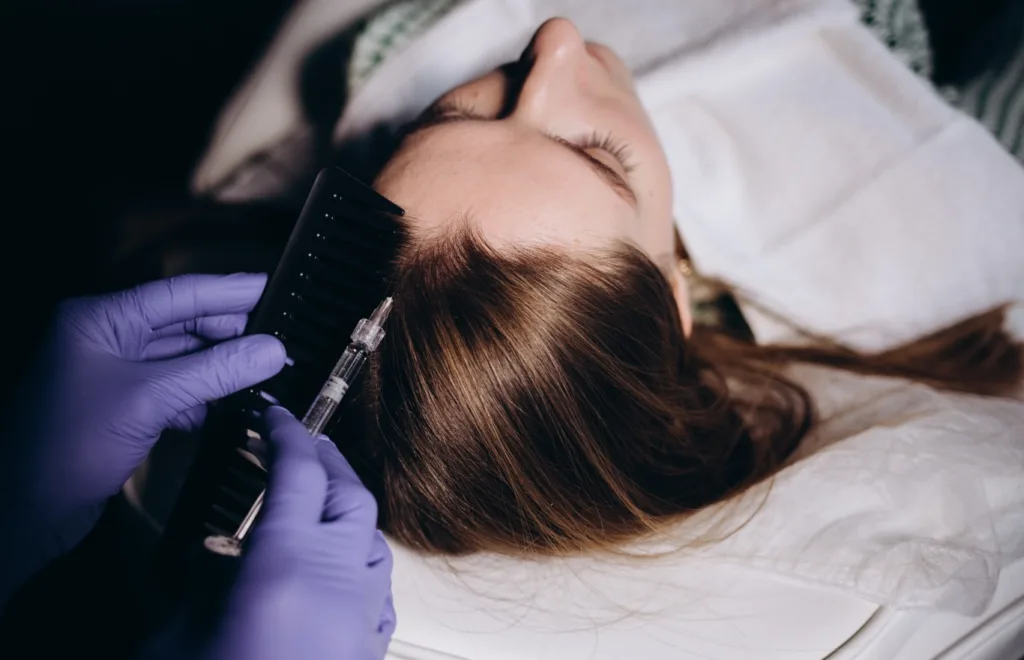 A high-angle close-up shows a person with long brown hair lying down, receiving a PRP Hair Treatment. Gloved hands are holding a fine needle and a black comb, meticulously parting the hair and injecting a substance into the scalp near the hairline.