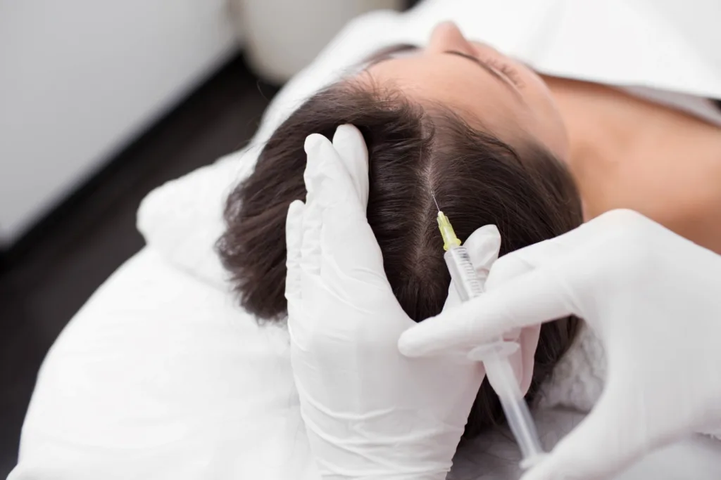 Close-up of a person receiving a cosmetic injection near their eye, possibly related to hair loss treatment in Novi (though not directly depicted in this specific image). A medical professional, wearing gloves, is holding a syringe and carefully administering the treatment, while the patient lies still with their eyes closed.
