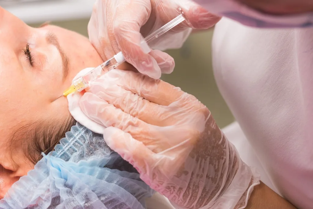 A close-up shot shows a medical professional in gloves administering an injection to a patient's face, highlighting potential Xeomin benefits for cosmetic concerns. The patient's eye is closed, and a cotton pad is held near the injection site, indicating a precise and careful procedure.