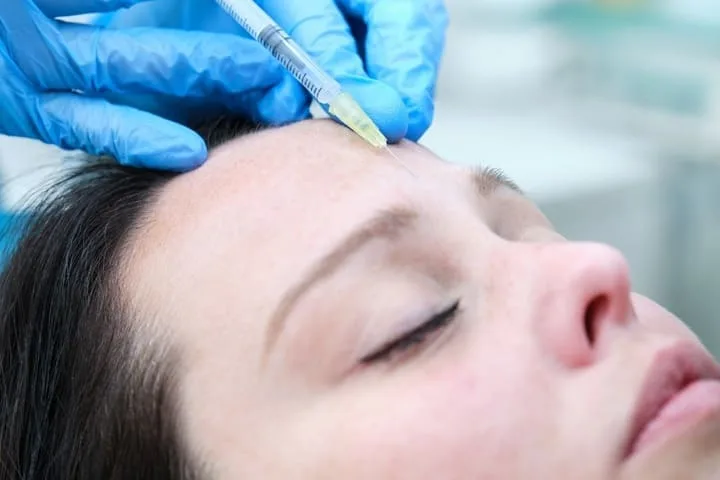 A close-up of a woman receiving anti wrinkle injections on her forehead by a professional wearing blue medical gloves